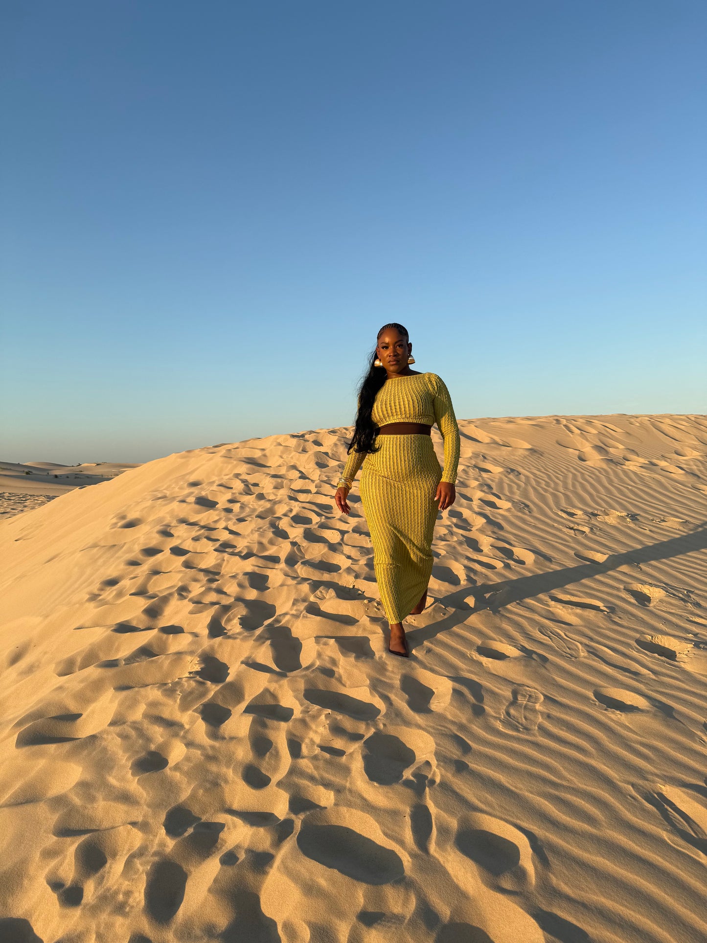 Model wearing the green Sahara Sands Two-Piece Set from YNAE B. Emerge Collection, featuring a knitted open-back, long-sleeve crop top with self-tie closure and a matching high-waisted skirt. Chic stretch-knit resort wear for destination events and summer getaways.